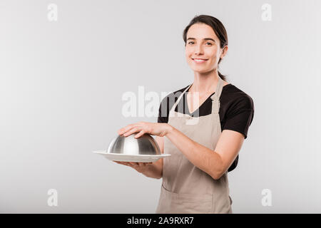 Happy young brunette waitress in apron keeping hand on cover of cloche Stock Photo