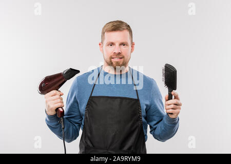 Happy young bearded hairdresser in apron holding hairdryer and hairbrush Stock Photo