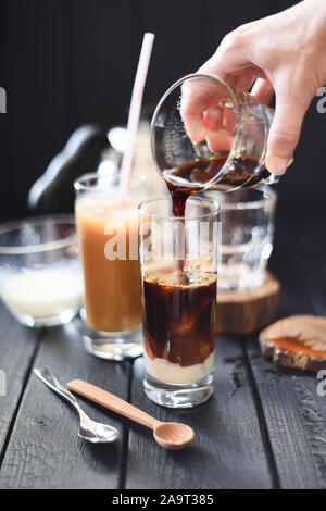 Making iced coffee in tall glasses. Woman hand pouring black coffee over ice cubes and condensed milk on black background selective focus Stock Photo