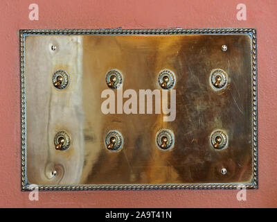 shiny brass switch-plate with eight old fashioned electrical switches on rusty ochre painted wall in historic house in England UK Stock Photo