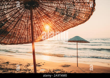 Umbrella On Sandy Beach. Sun Sunshine Through Wicker Umbrella At Summer Sunrise Sunset. Stock Photo