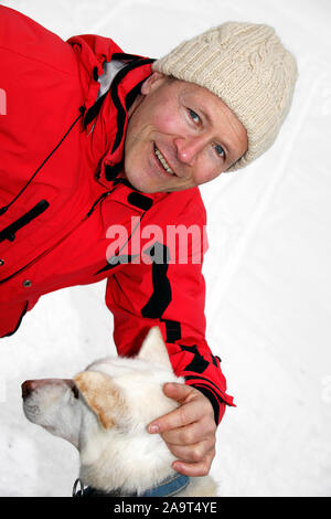 Junger Mann in roter Jacke streichelt Sibirischen Husky / Canis lupus familiaris im Schnee, Schlittenhunde im Winter, MODEL RELEASE VORHANDEN Stock Photo