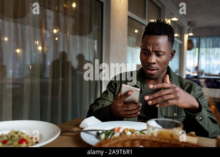 young disappointed surprised african american man messaging online, checking social networks in cafe. copy space Stock Photo
