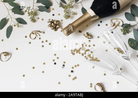 Happy New Year composition. Champagne glasses nad wine bottle with golden confetti stars and eucalyptus branches isolated on white table background Stock Photo