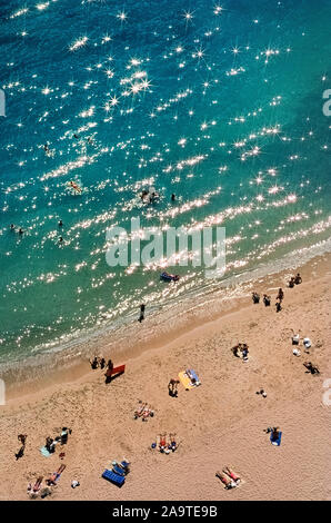 Sunlight sparkles off the Pacific Ocean at at the edge of famous Waikiki Beach, a popular destination for vacationers who come to the capital city of Honolulu on Oahu, one of the eight major islands that are part of the tropical state of Hawaii, USA. This aerial photograph shows only a small part of  Waikiki Beach, which is two miles (3.2 kilometers) long and actually has eight sections with individual beach names. More populated are the sands and walking promenades in front of historic and high-rise hotels along the meandering shoreline. Stock Photo