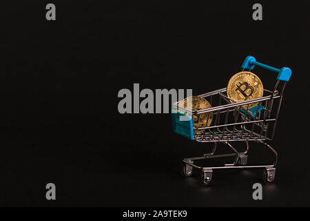 Gold bitcoin coins sparkle in a supermarket trolley on a black background. Concept for financial articles and forecasts. Stock Photo