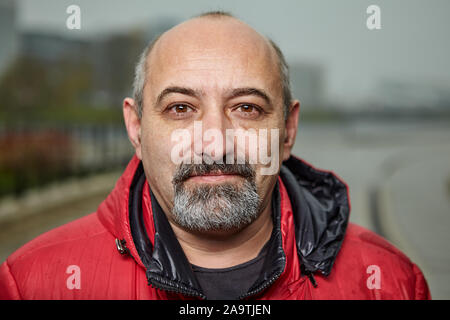Close up portrait of a bald man with a gray beard outdoors in cloudy weather. Mature adult male in a red synthetic jacket, stands on a city street. Bu Stock Photo