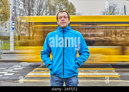 One middle-aged man, about 50, is standing on a city street, opposite a pedestrian crossing and yellow regular bus passing by. A sad male of fifty yea Stock Photo