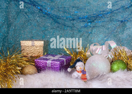 A smiling snowman on a white fur base next to Christmas balls and gift boxes. Blue background with the effect of falling snow. Selective focus. Stock Photo