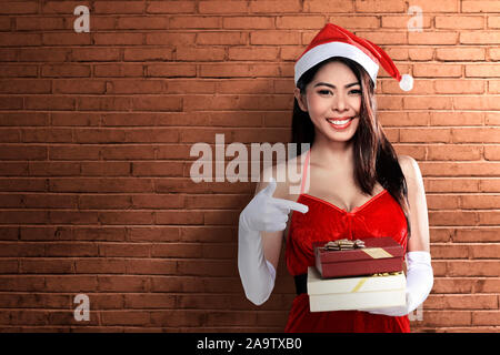 Asian woman in Santa costume holding gift box with brick wall background Stock Photo