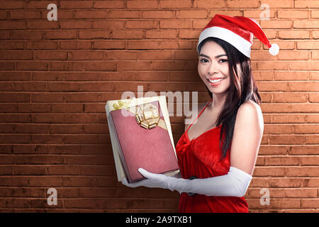 Asian woman in Santa costume holding gift box with brick wall background Stock Photo