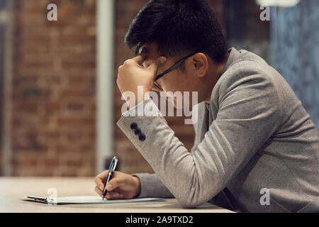 Side view portrait of young Asian man writing on clipboard while working at desk in modern office, copy space Stock Photo