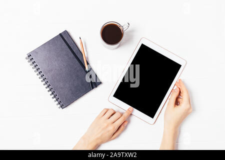 Female hands using tablet computer with blank display next to cup of coffee and paper notebook on white desktop, flat lay Stock Photo
