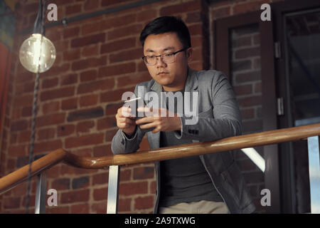 Low angle portrait of young Asian businessman using smartphone standing on balcony in modern office, copy space Stock Photo