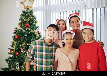 Portrait of hugging smiling Vietnamese family of three generations standing at room with Christmas tree Stock Photo