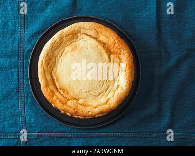 Plate with whole cheesecake pie on table with jeans tablecloth. Classic cheesecake flat lay or top view. Vertical. Stock Photo