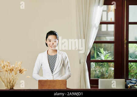 Young beautiful Vietnamese woman in white jacket working on laptop at hotel reception Stock Photo