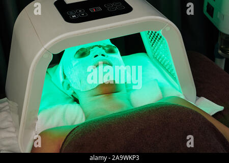 Young woman with sheet mask on her face having LED light treatment in beauty salon Stock Photo