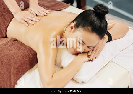 Smiling pretty young Asian woman lying on bed in spa salon and enjoying nice relaxing massage Stock Photo