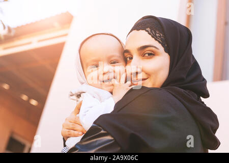 Young Muslim woman in hijab holding her 1-year-old baby boy, hugging and kissing. Happy Muslim mother in abaya clothing kiss and play with her little Stock Photo