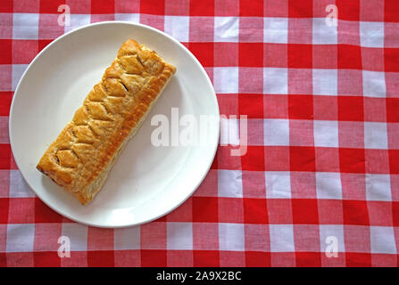 Flat lay of sausage roll on a plain white side plate on a red checked background with white space Stock Photo