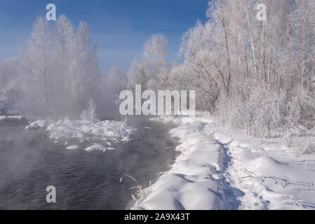 Amazing winter landscape with birch trees and bushes in hoarfrost, a path in the snow, a river and fog above the water against a blue sky in severe fr Stock Photo