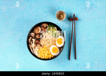 Ramen. Soba noodles with eggs, shiitake mushrooms, and vegetables, top shot on a blue background with chopsticks and a cup of sake Stock Photo