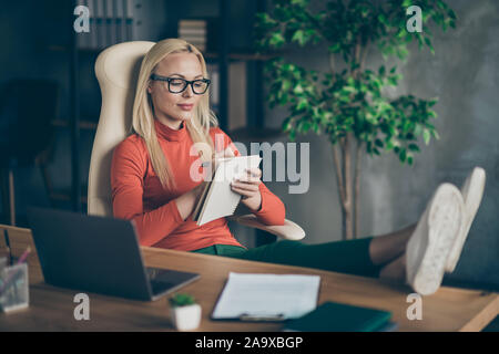 Photo of serious confident woman looking into notebook writing down her thoughts needed to be used in start up with feet on desktop Stock Photo