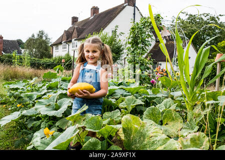 Girl standing in a vegetable patch in a garden, holding yellow gourd, smiling at camera. Stock Photo