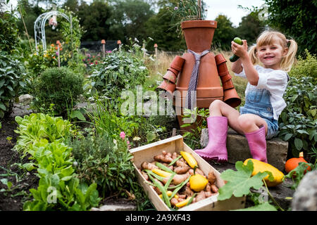 Smiling blond girl sitting in garden next to terracotta scarecrow, picking fresh vegetables. Stock Photo