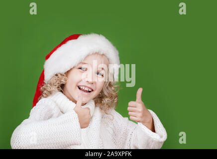 Merry Christmas Little girl in Santa hat showing thumb up on bright green vivid color background. Red and green. Portrait of lovely elf child. Copyspa Stock Photo