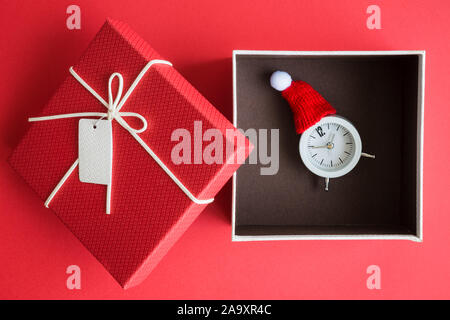 Flat lay of small alarm clock wearing red winter hat in gift box against red background creative christmas holiday present concept. Stock Photo