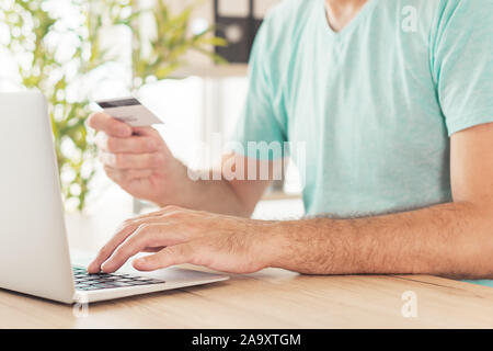 Online shopping with credit card and laptop computer, casual man is using modern technology in home office to purchase items on internet, close up of Stock Photo