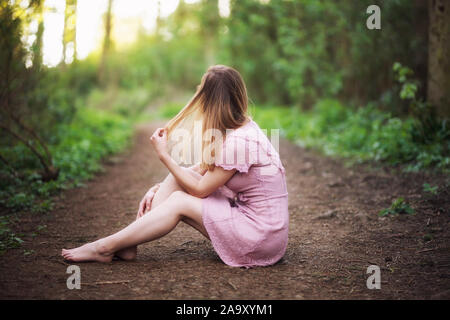 Young woman in pink dress sitting in forest meadow road and touch her hair Stock Photo