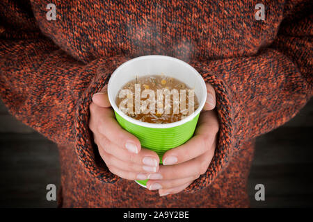 Hot tea in hands. Close up of woman hands holding a paper cup with herbal tea. Focus on hands and cup. Stock Photo