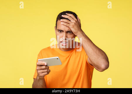Bothered and upset shocked young handsome man, holding smartphone horizontally watch devastating news or video, punch forehead, frowning, lost in game Stock Photo