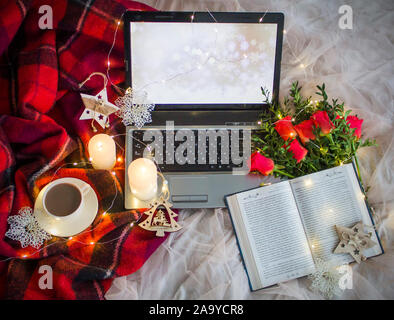 Composition: a laptop, a cup of coffee (tea), red roses with boxwood, a book, candles, a garlands and a red blanket. Stock Photo