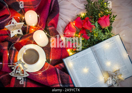 Composition: a cup of coffee (tea), red roses with boxwood, a book, candles, a garlands and a red blanket. Stock Photo