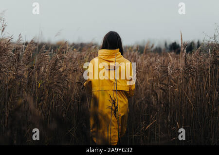 Female in high grass wearing yellow rain coat and looking away from camera - Moody fall scenery with a young girl in bright clothing walking in high g Stock Photo