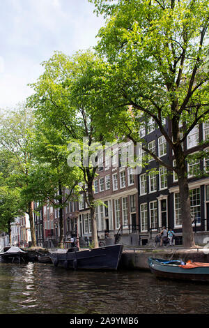 View of canal, woman riding bicycle, parked boats, trees and historical, traditional buildings showing Dutch architectural style in Amsterdam. It is a Stock Photo