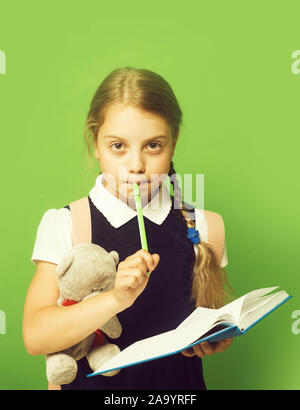 Study and back to school concept. Kid in school uniform isolated on green background. Pupil holds blue book, marker and teddy bear. Girl with braids and thoughtful face expression Stock Photo