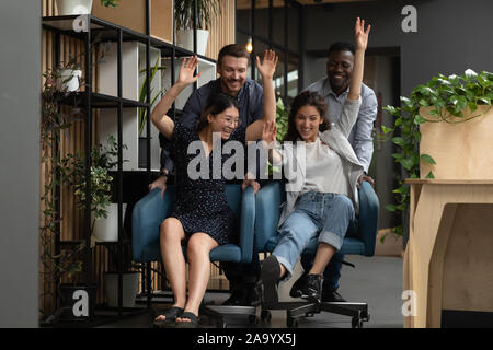 Overjoyed young diverse colleagues having fun, riding on chairs. Stock Photo