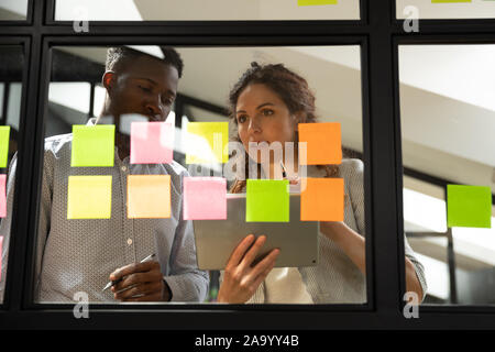 Concentrated mixed race young colleagues standing near glass kanban board. Stock Photo