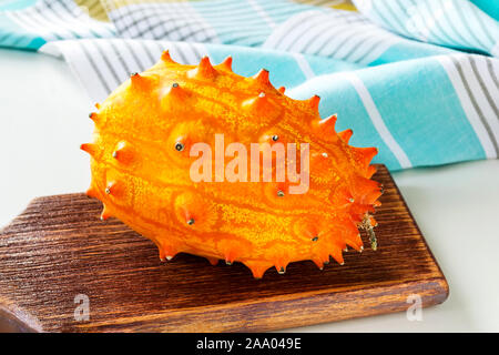Whole fresh orange kiwano (cucumis metuliferus, horned melon) on a brown wooden cutting board over kitchen table. Fruits, vegetables, vegetarian. Stock Photo