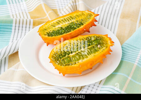 Two halves of fresh orange kiwano (cucumis metuliferus, horned melon) on a white plate over a table. Fruits, vegetables, vegetarian and healthy eating Stock Photo