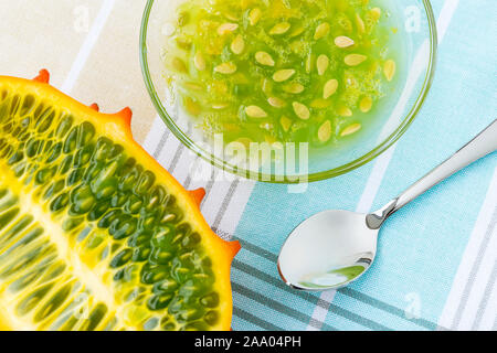 Half of orange kiwano (cucumis metuliferus, horned melon), glass bowl with kiwano pulp and a spoon on a tablecloth. Fruits, vegetables, vegetarian. Stock Photo