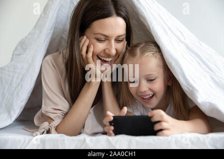 Happy woman lying under blanket with daughter, playing mobile games. Stock Photo