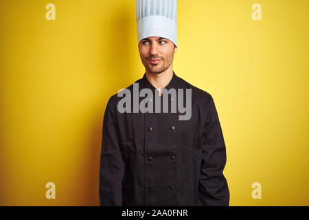 Young chef man wearing uniform and hat standing over isolated yellow background smiling looking to the side and staring away thinking. Stock Photo