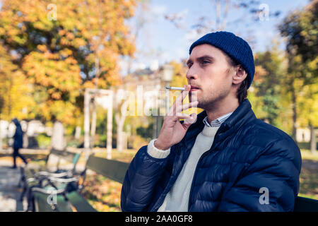 Young man in a cap smoking a cigarette on a bench during a nice autumn day. Stock Photo