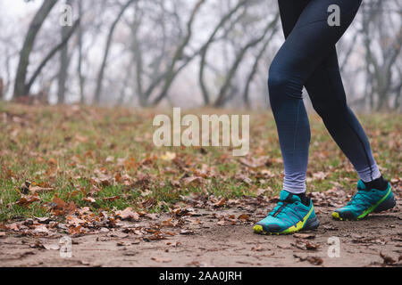 Close up of young fitness woman legs running at forest trail. Fallen foliage on the ground, autumn foggy morning. Shooting point from below. Stock Photo